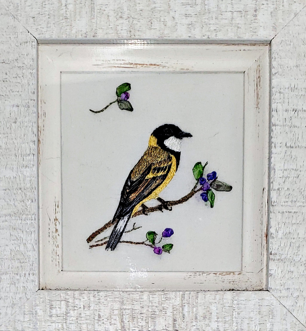 Embroidered picture of a bird on a branch with berries in a white frame.